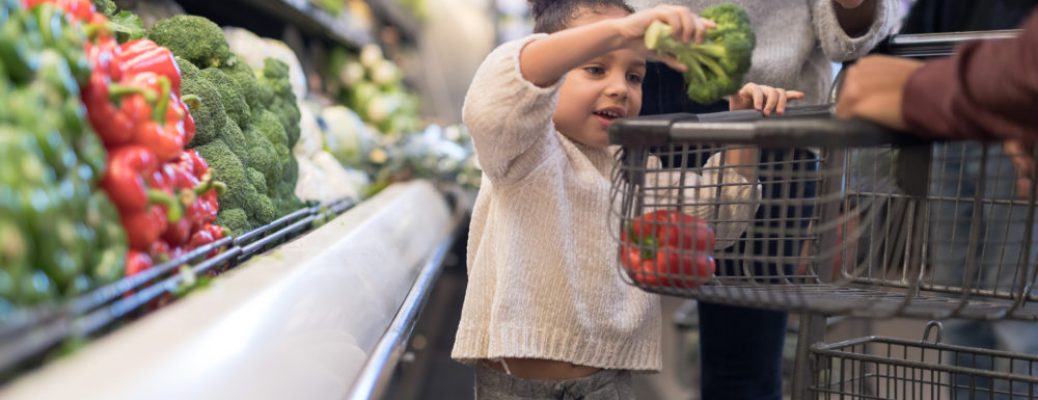 A pre-school age girl helps her parents pick out veggies in the produce section at the grocery store. She is putting a head of broccoli in the cart.